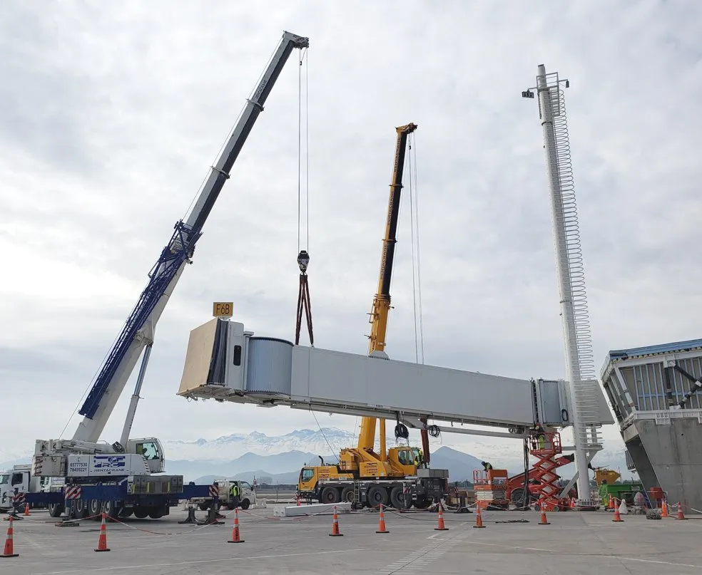 Dos grúas izando el túnel de una pasarela de embarque durante su instalación, con la cordillera al fondo